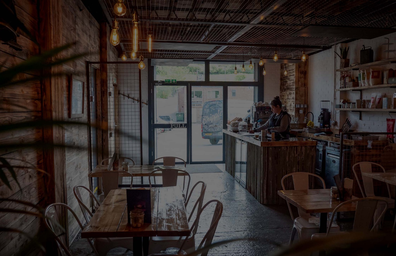 View of a coffee shop with an out of focus plant, tables with menus on and a counter with a lady making coffee behind.